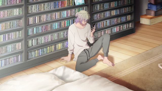 Shin Iryuu from A Sign of Affection sits on a wooden floor leaning against a large shelf filled with CDs. He holds a smartphone in one hand while looking at it with a calm, slightly pensive expression. He wears a loose white long-sleeve shirt and grey sweatpants, barefoot, in a softly lit room with a futon visible nearby.