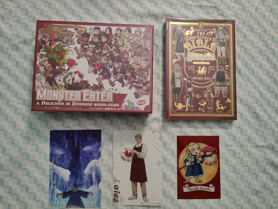 A photo with some items from the DunMeshi Exhibit merch store laid out: The Dungeon Meshi Board Game, the Complete Adventurer's Bible by Ryoko Kui, a postcard of Falin in the ice level, a postcard of Laios in his cafe outfit, and a clear card of chibi Marcille